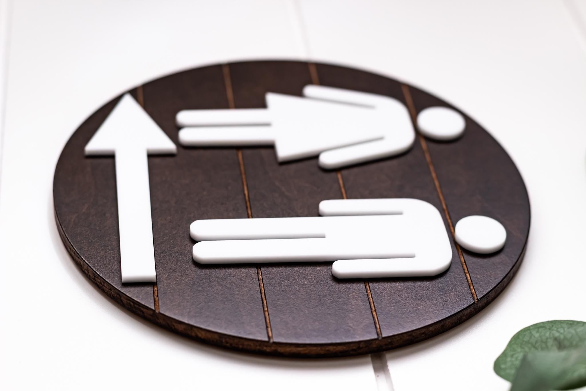 Directional restroom sign with white female and male icons and right-pointing arrow on dark round backing