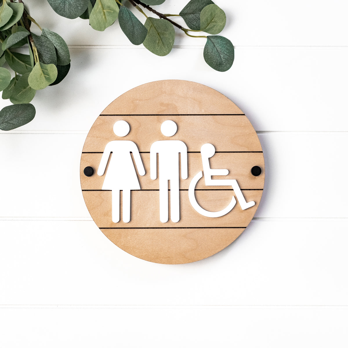 Round 3D restroom accessibility sign on light wood with wheelchair symbol and gender icons
