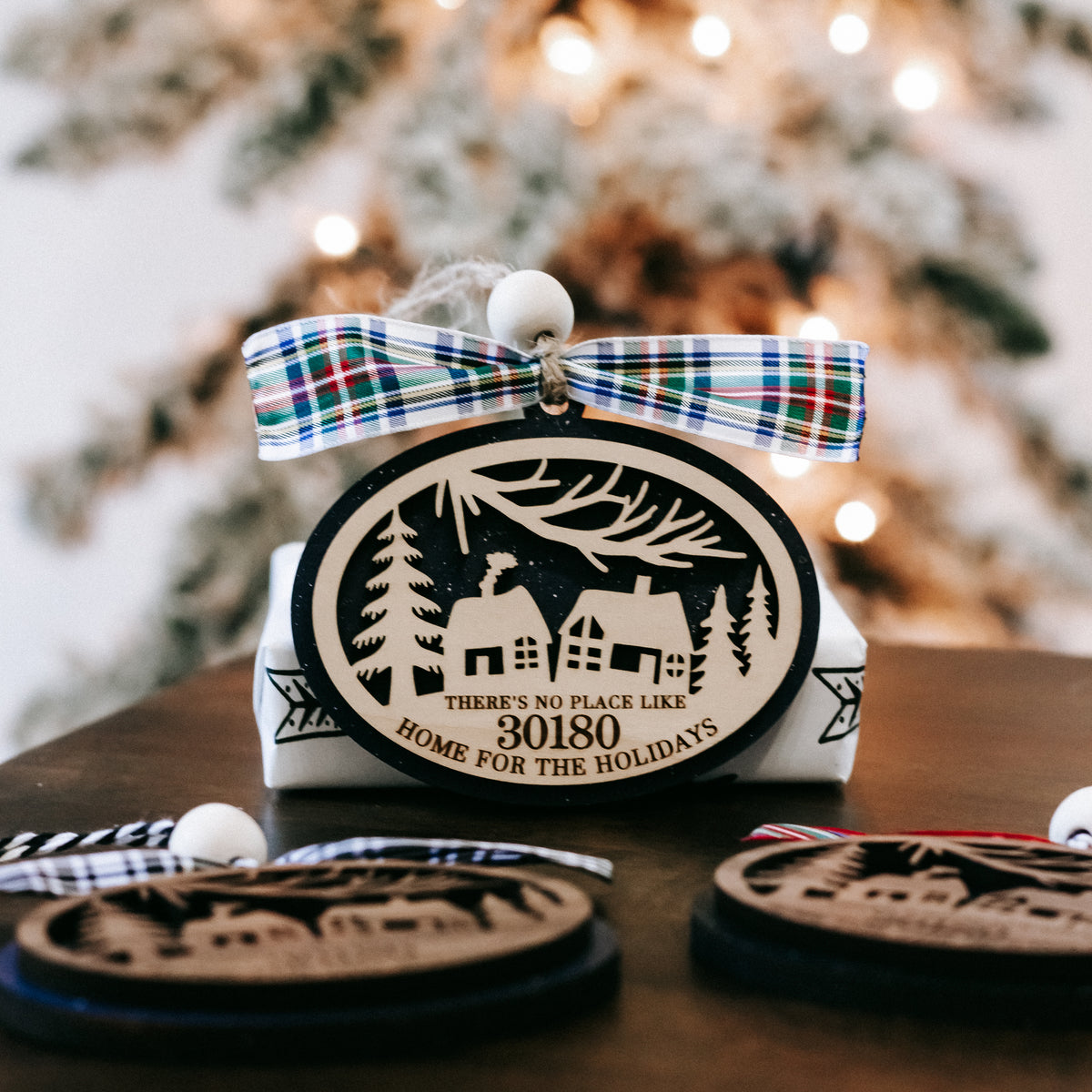 Round wood Christmas ornament in box with blue plaid bow and house scene with pine trees