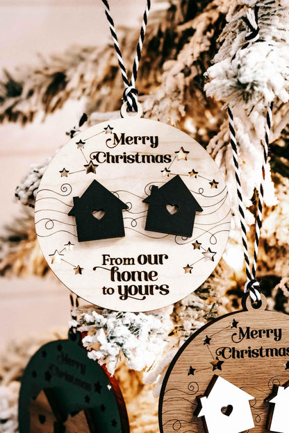 Hanging round wooden Christmas ornament with two black house icons and “From our home”