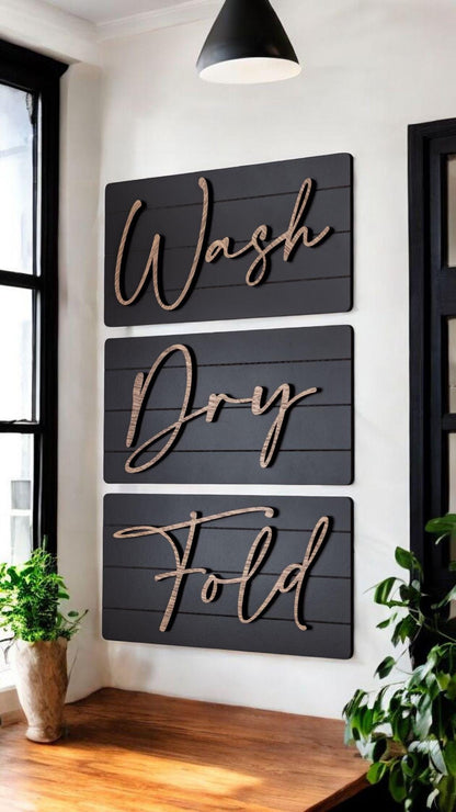 Three dark wall signs reading Wash, Dry, Fold with wood-tone cursive lettering on wall