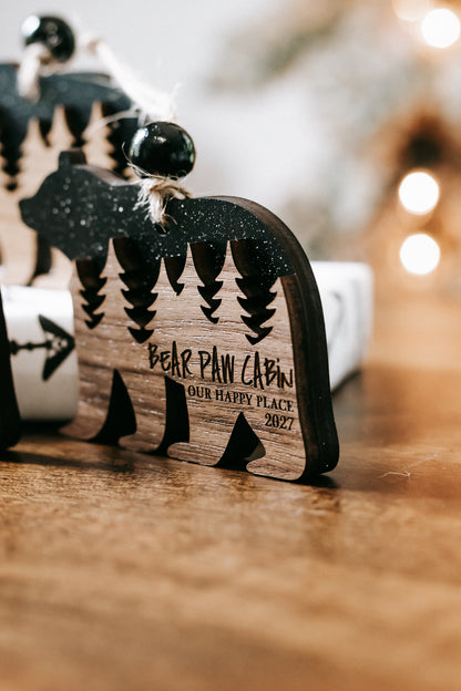 Bear cabin ornament close-up with dark glitter bear, engraved pine trees, and text