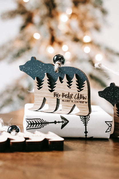Bear cabin ornament with dark glitter finish, engraved pine trees, and cabin coordinates