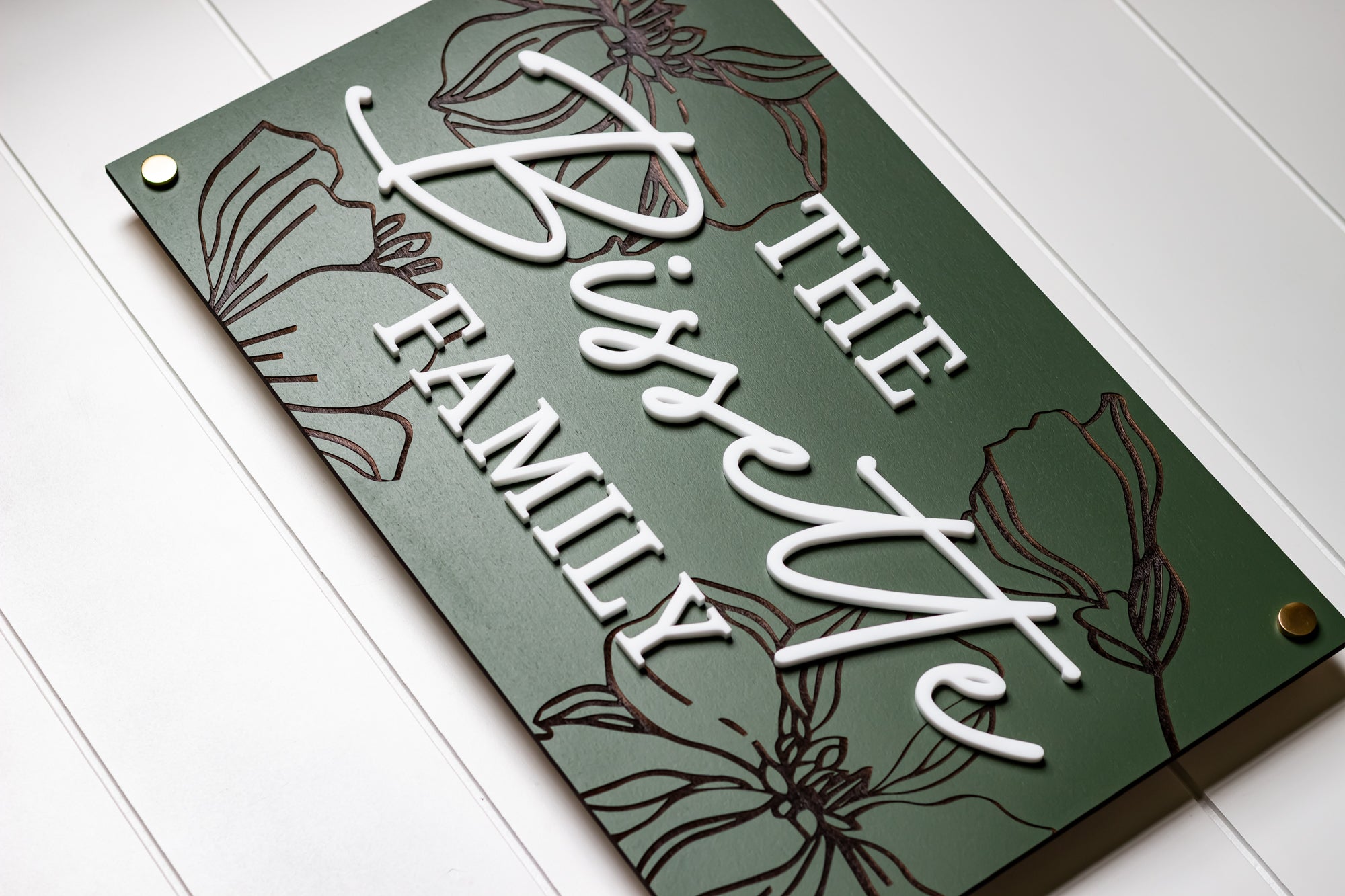 Green rectangle family name sign with white engraved script, flowers, and bronze fasteners
