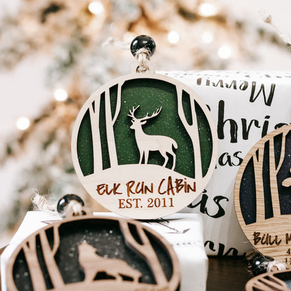 Hanging round wooden Christmas ornament with green deer design and “Elk Run Cabin” text