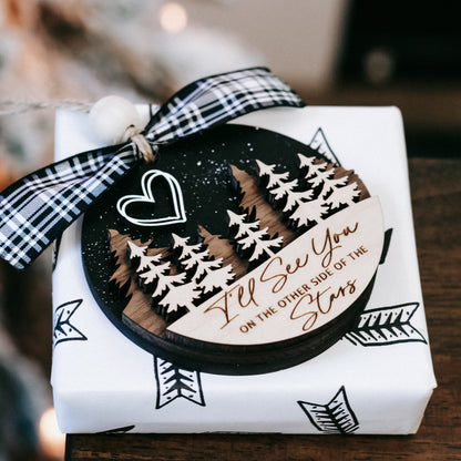Wrapped memorial Christmas ornament with plaid bow, heart icon, and pine trees on box.