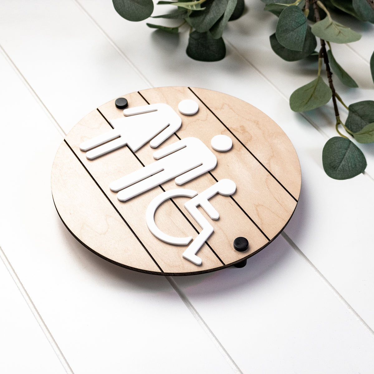Round 3D accessibility restroom sign with raised wheelchair icon in white on light wood