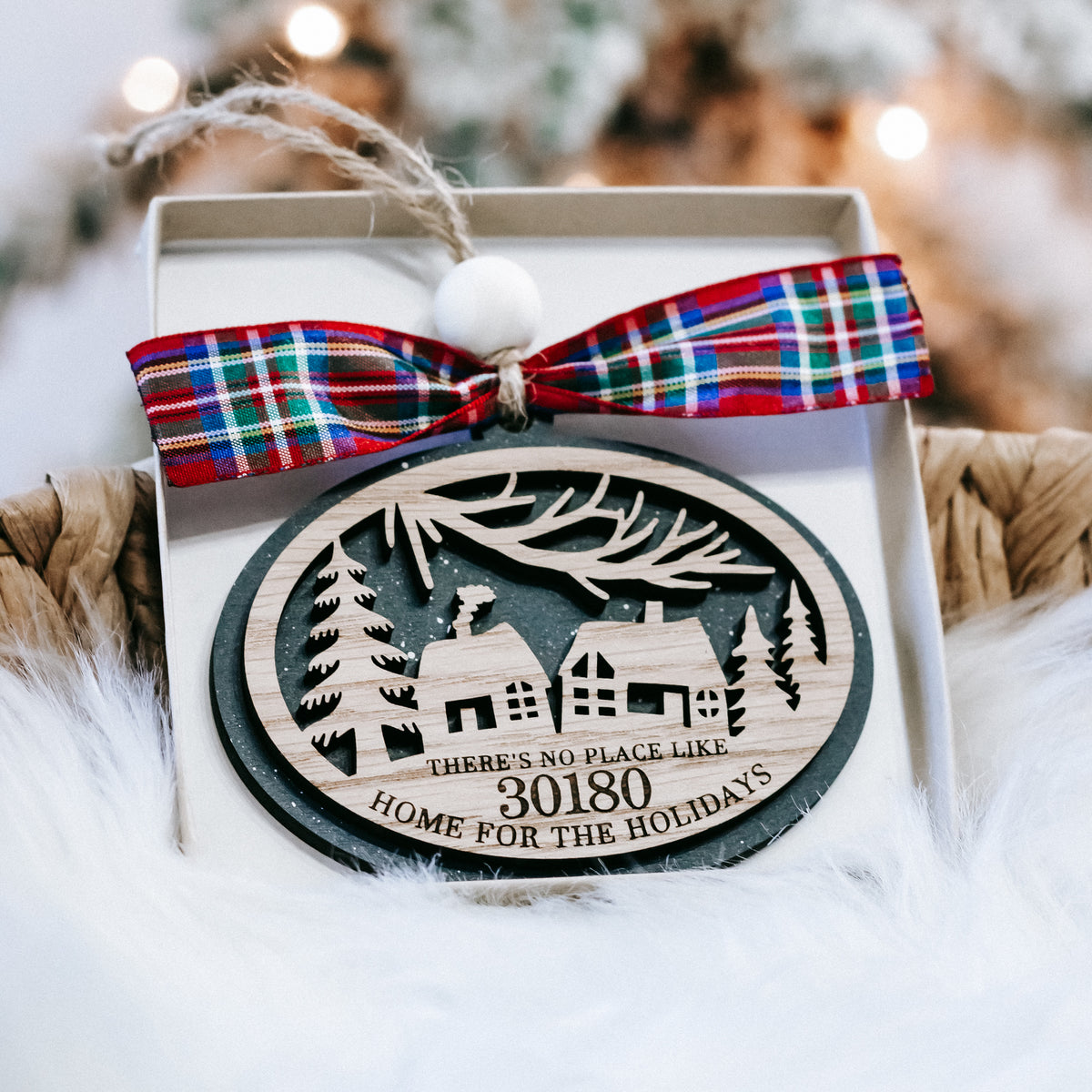 Round personalized Christmas ornament in gift box with plaid bow and house-and-tree scene