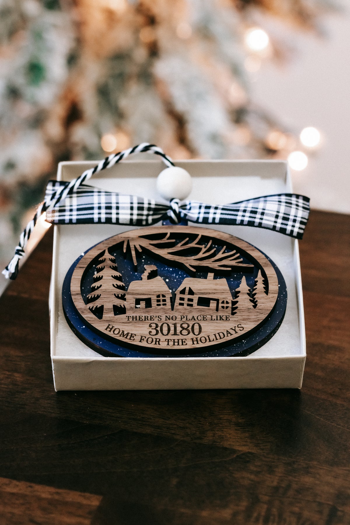 Round wood Christmas ornament in box with black plaid bow and house-and-trees design